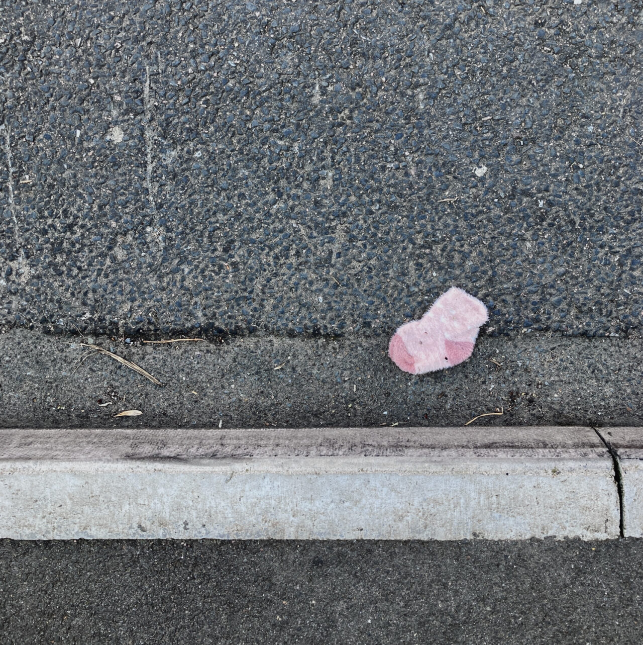 Photograph looking down at a pink baby's sock in a gutter by the side of the road, with pavement at the bottom.