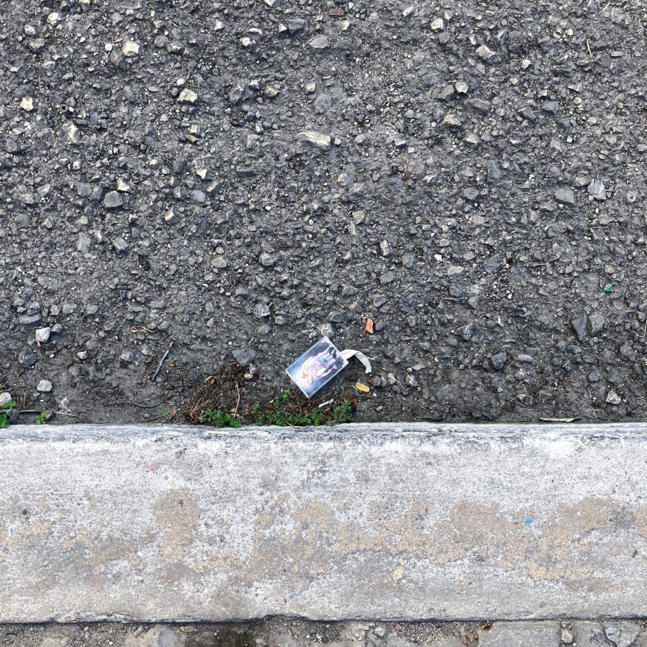 Photograph looking down at a prayer card in a gutter by the side of the road, with pavement at the bottom and road above.