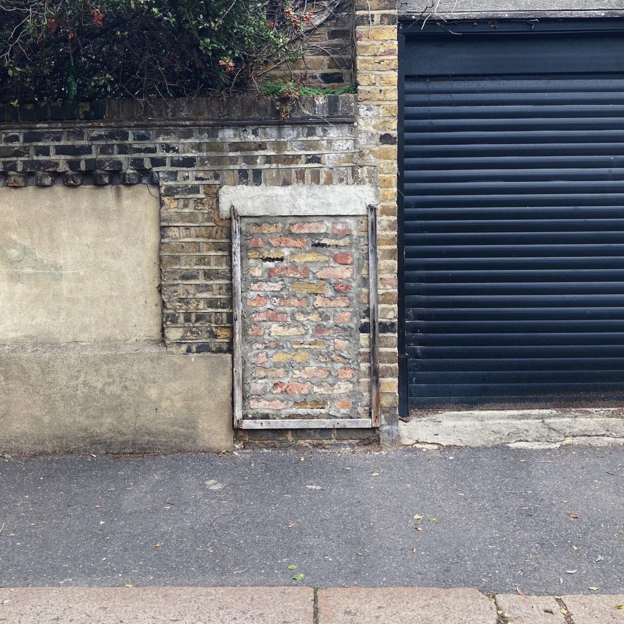 Walthamstow. Photograph of a wall with the remains of a frame that used to hold a notice, with a black garage door to the right and pavement in front