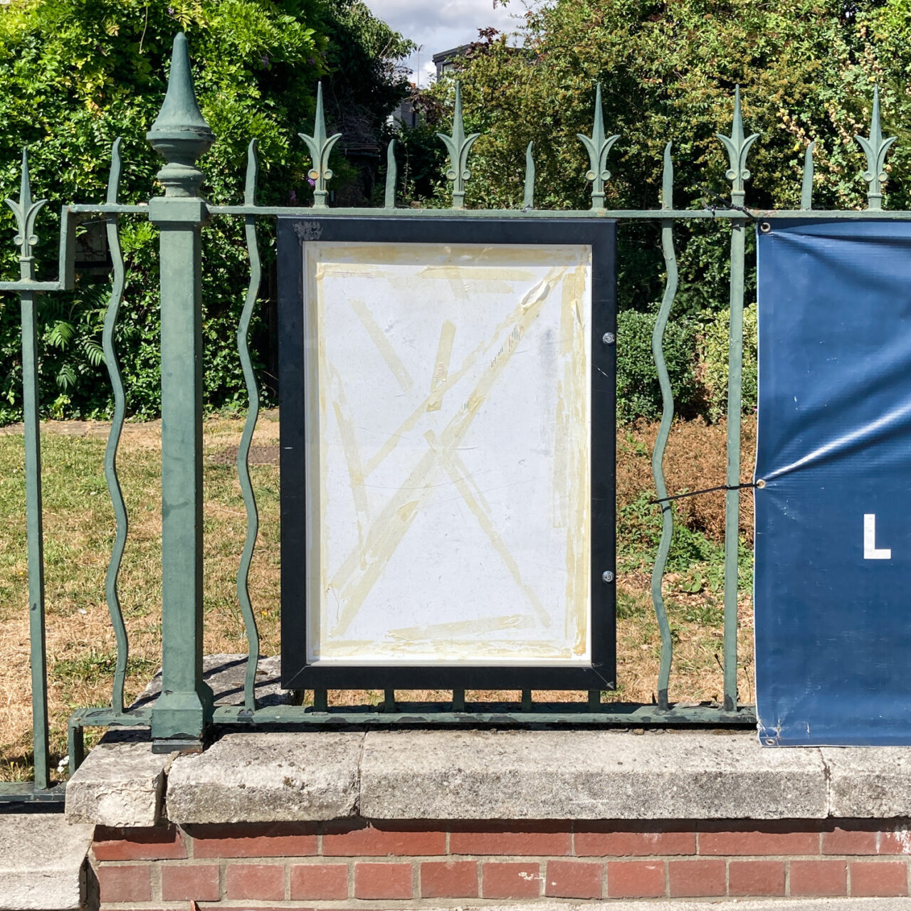 Walthamstow. Photograph of a black frame with white paper and tape marks inside, on an ornate green railing with trees behind and brick wall below.