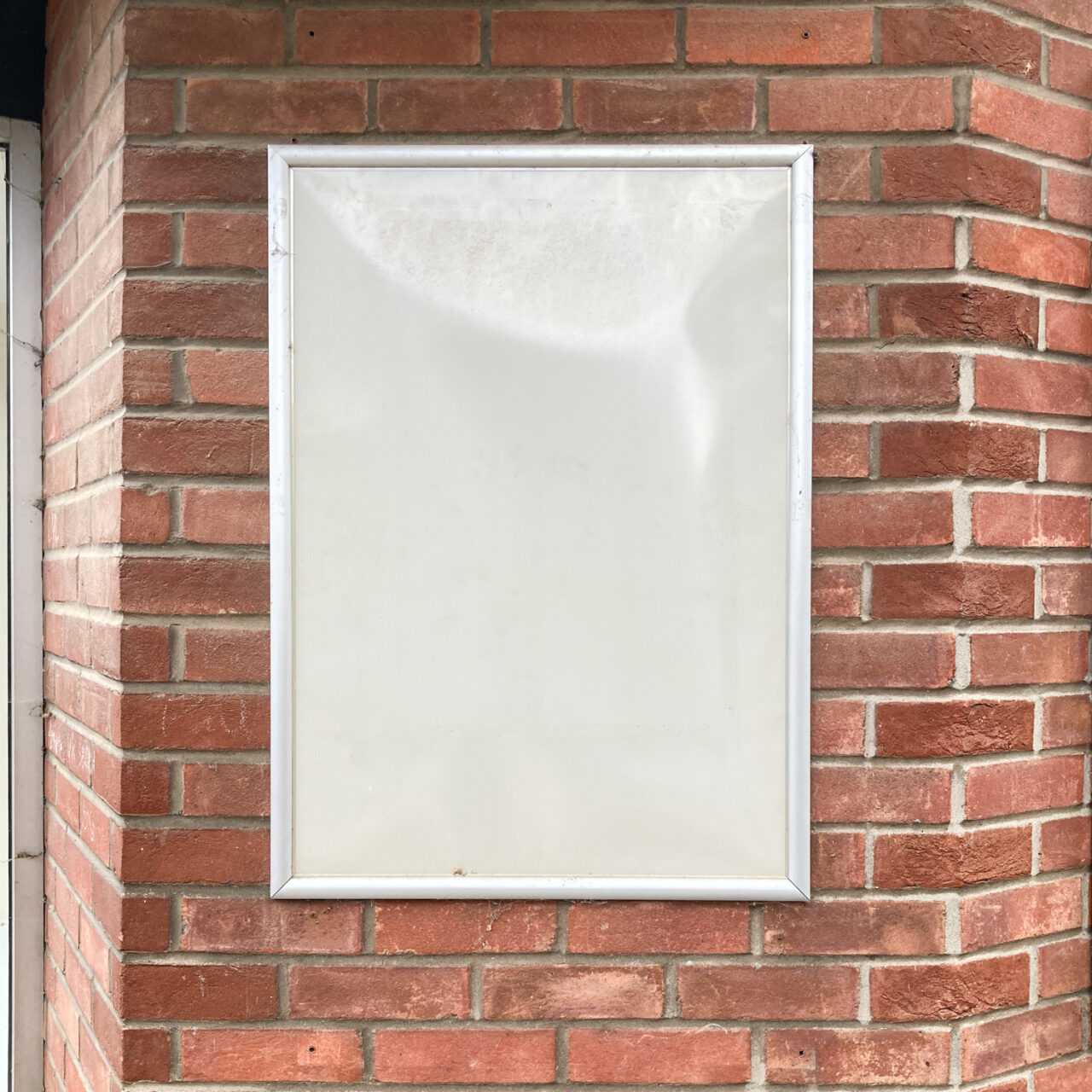 Wivenhoe. Photograph of an empty metal-framed noticeboard on a brick wall.