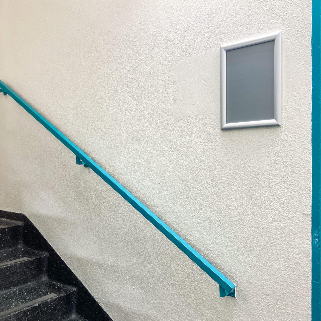 Gatwick. Photograph of a grey-filled empty silver frame on a wall, with stairs and a metal handrail below and to the left.