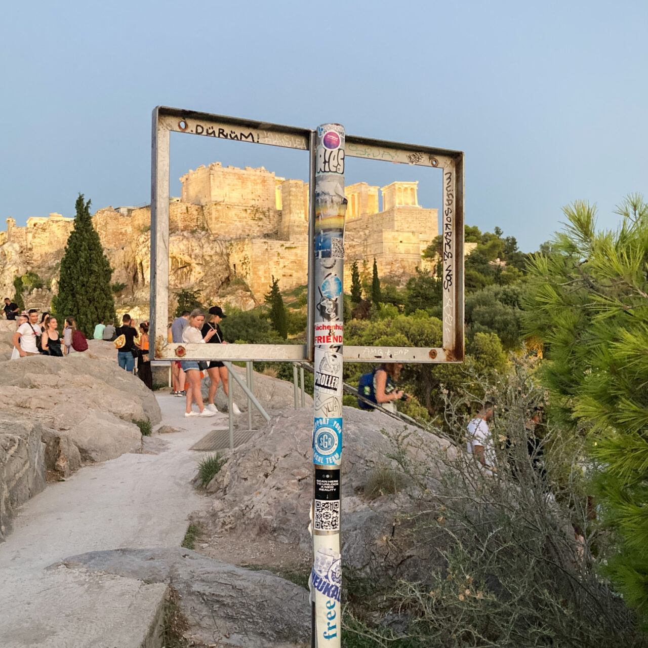 Athens. Photograph of an empty metal noticeboard with the Coliseum visible through it behind, with people on a rocky mound in the foreground.