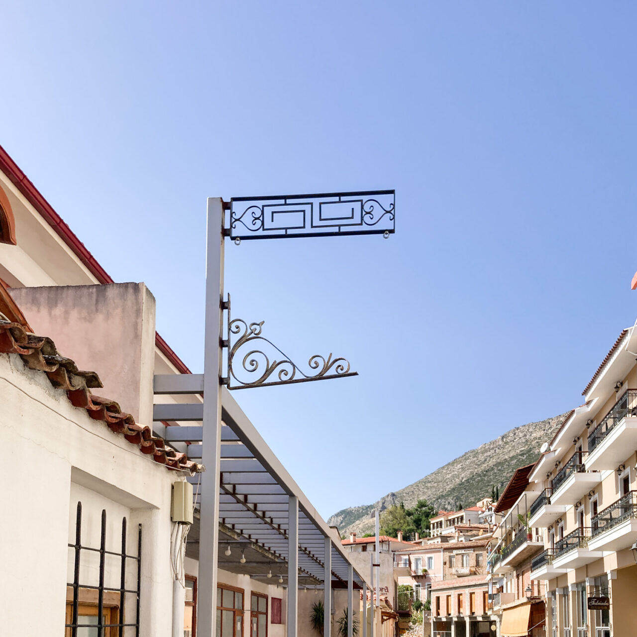 Delphi. Photograph of an ornate empty signpost against a blue sky, with buildings and mountain below and behind.