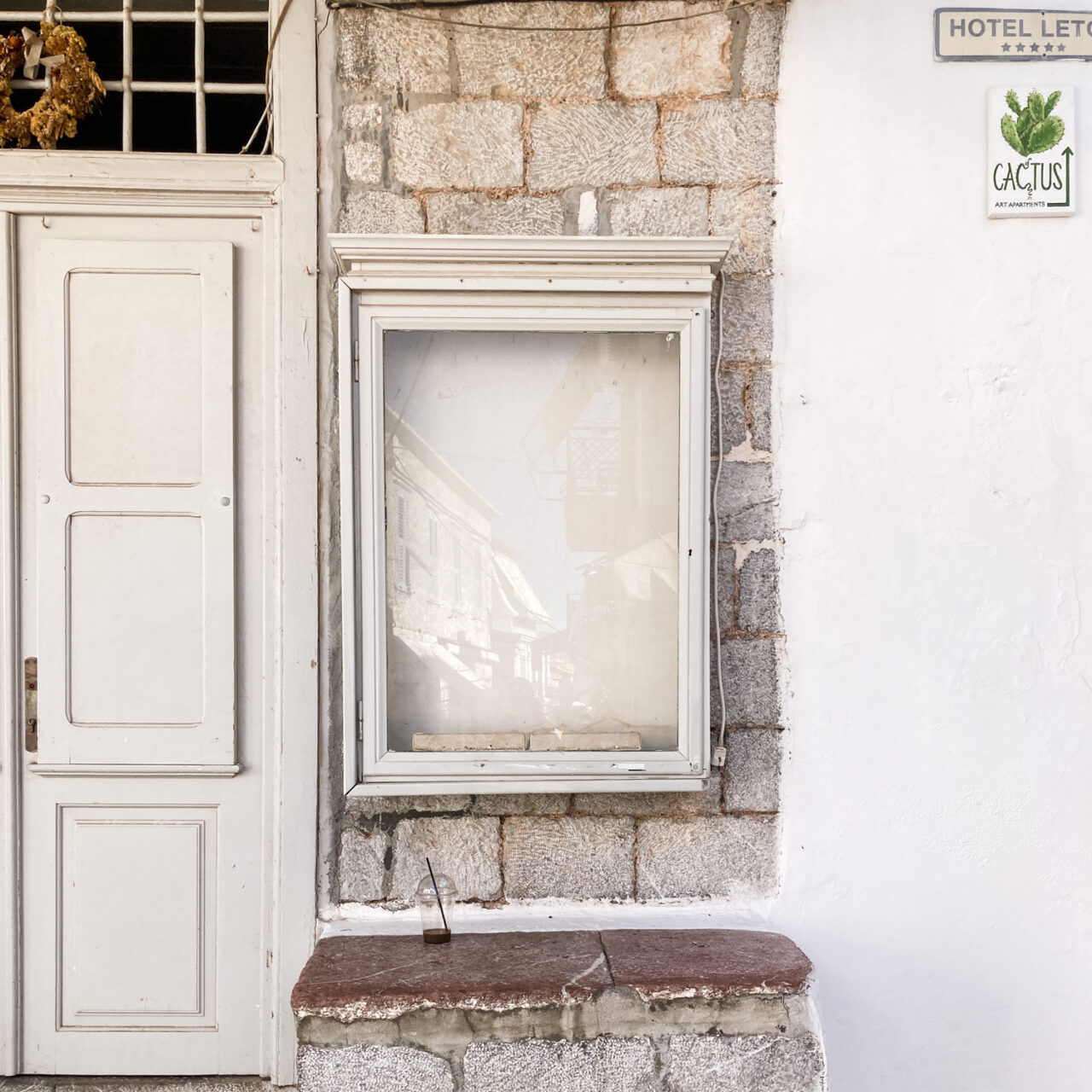 Hydra. Photograph of an empty noticeboard on a wall, with a grey door to the left and a white all to the right with signs reading 'Hotel Leto' and 'Cactus' top right.