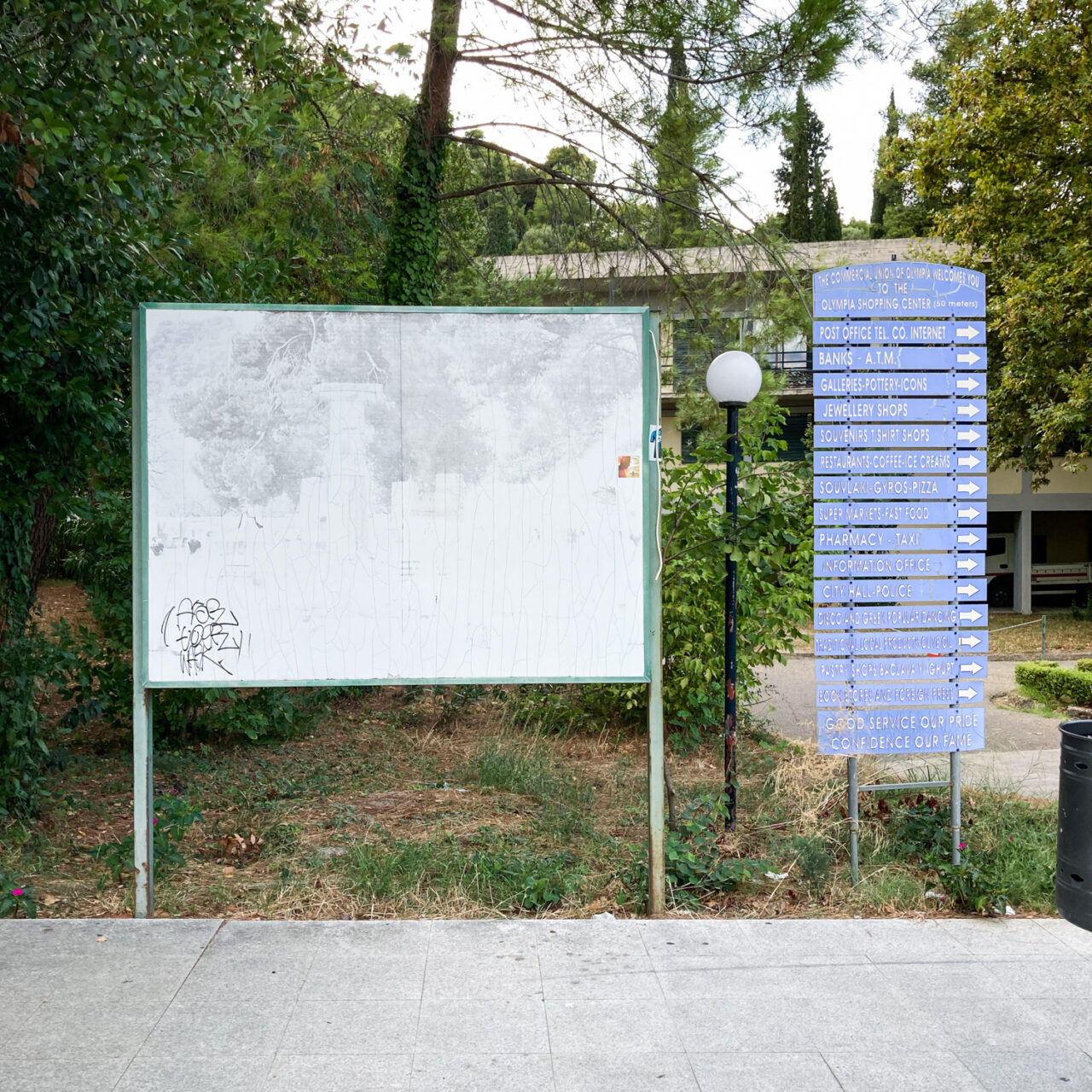 Ancient Olympia. Photograph of a board with a really faded image of columns just visible, with a lampost and a signpost with multiple destinations and arrows to the right, trees behind and a paved flow below.