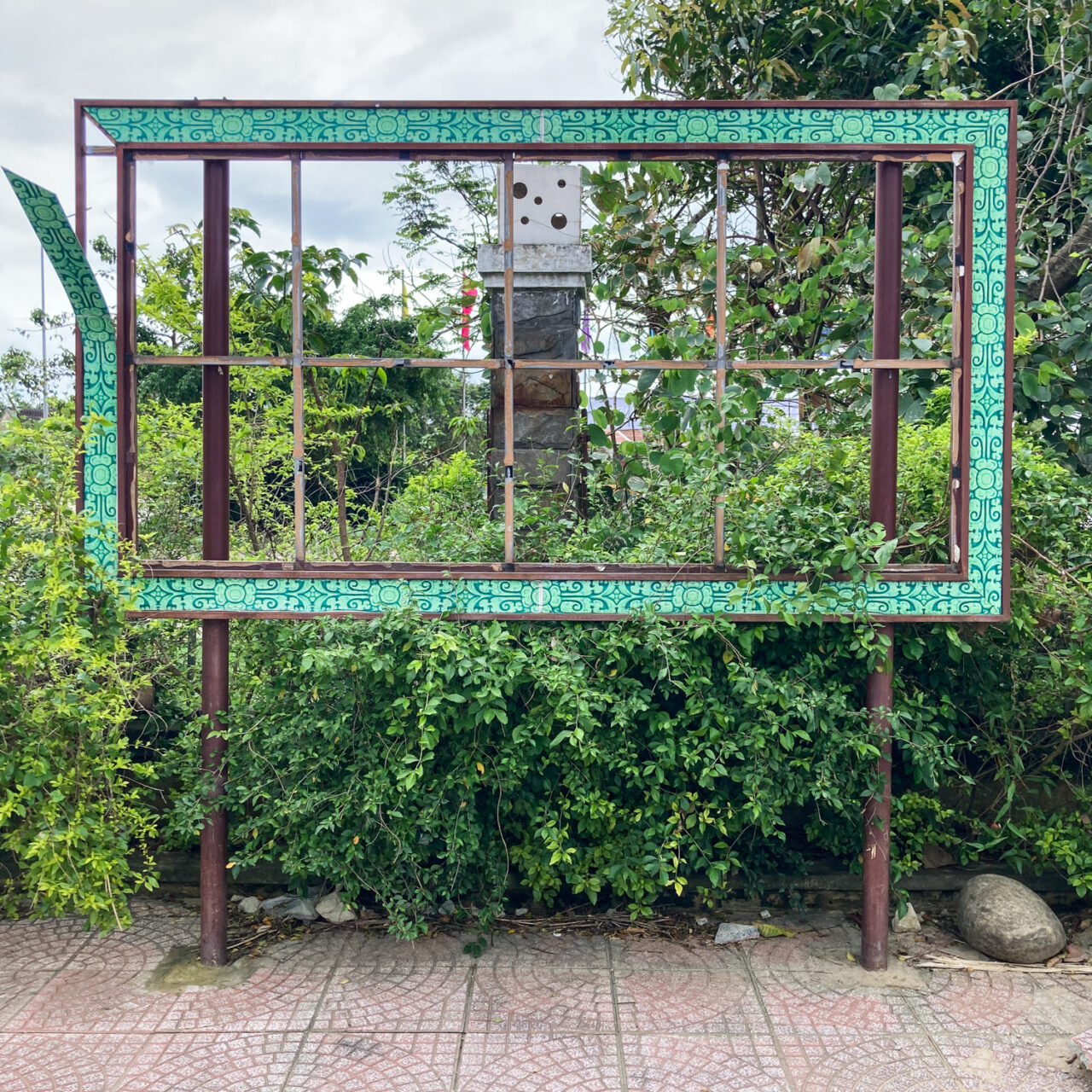Photograph of an empty metal noticeboard with an ornate green border with plants behind and pavement in the foreground.