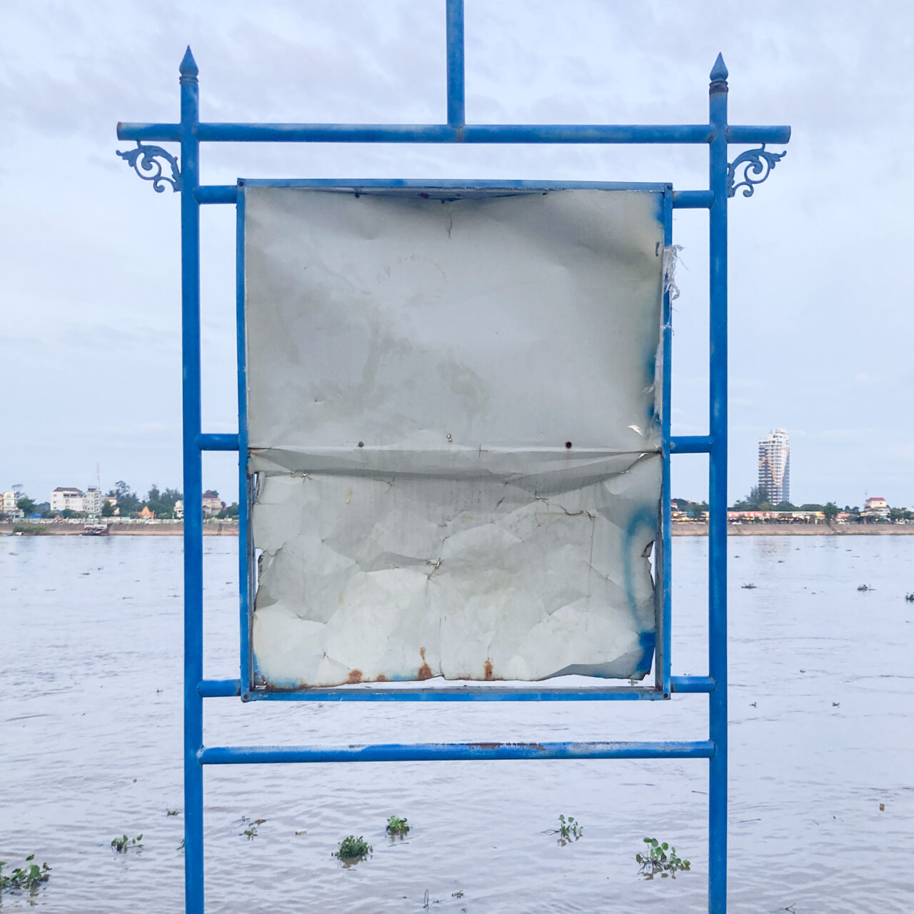Photograph of an empty metal blue noticeboard with a wide river behind and buildings visible on the land beyond it.