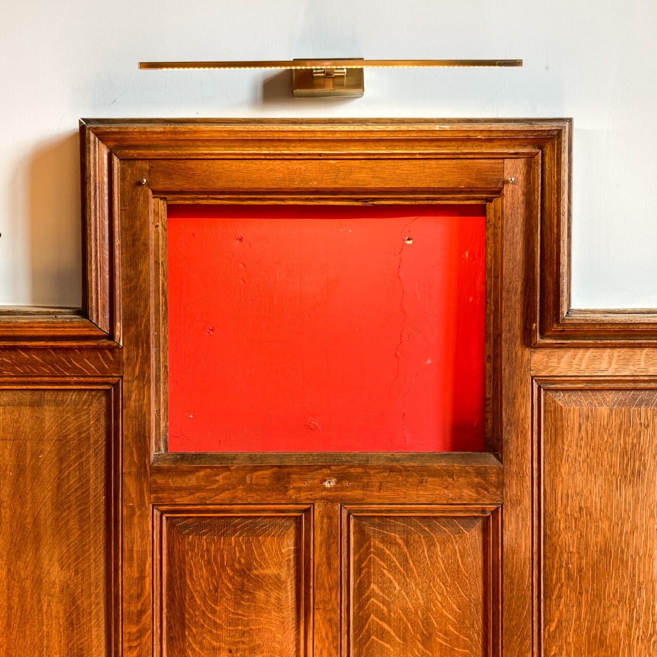 Photograph of a red display area lit by a wall lamp above it, set in wooden panels in the wall.