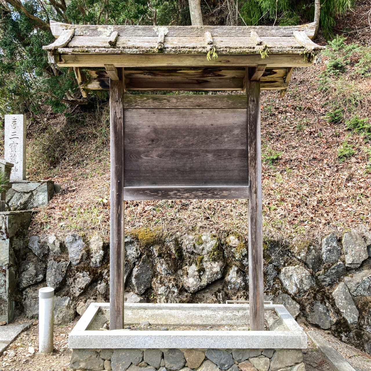 Photograph of an empty wooden noticeboard with a traditional Japanese roof above it, standing in a stone platform, with a stone wall and hill behind it.