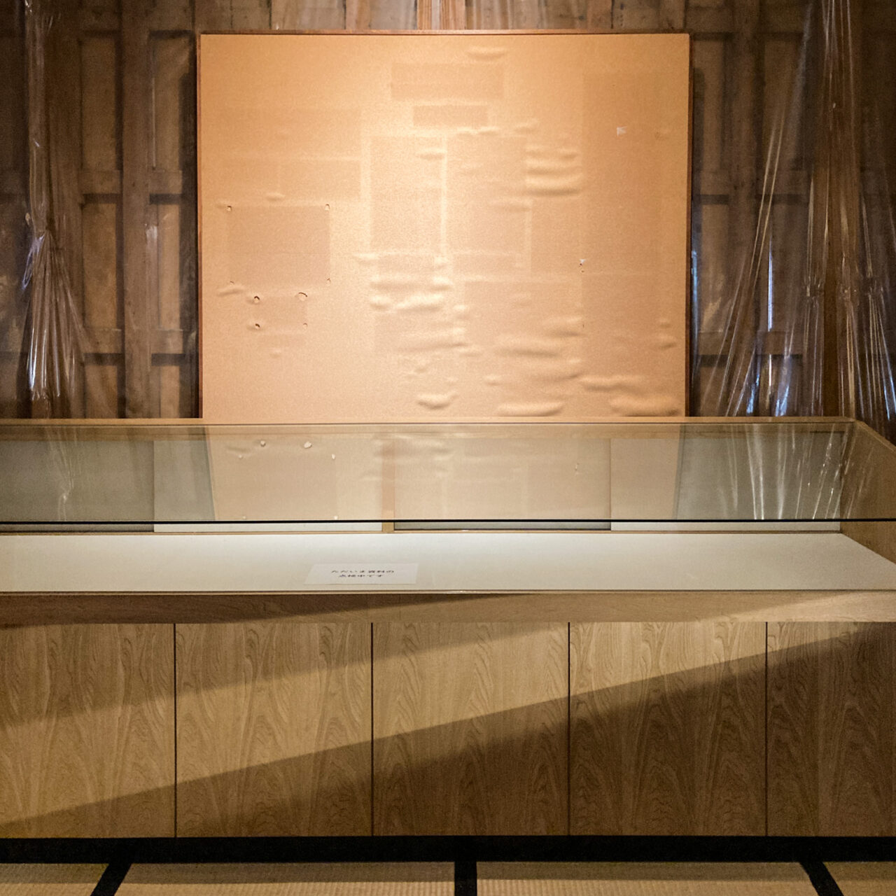 Photograph of an empty noticeboard with marks showing where the fabric has faded around the display that was previously mounted there. Behind is a wooden wall covered in plastic sheeting and in front is an empty display case.