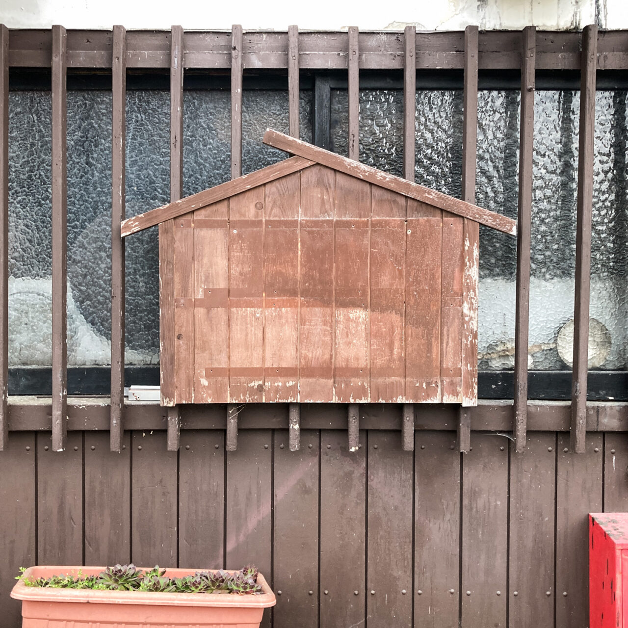 Photograph of an empty wooden noticeboard in the shape of a simple house, mounted on wooden bars in front of a window, with a wooden wall below and a window box bottom left.
