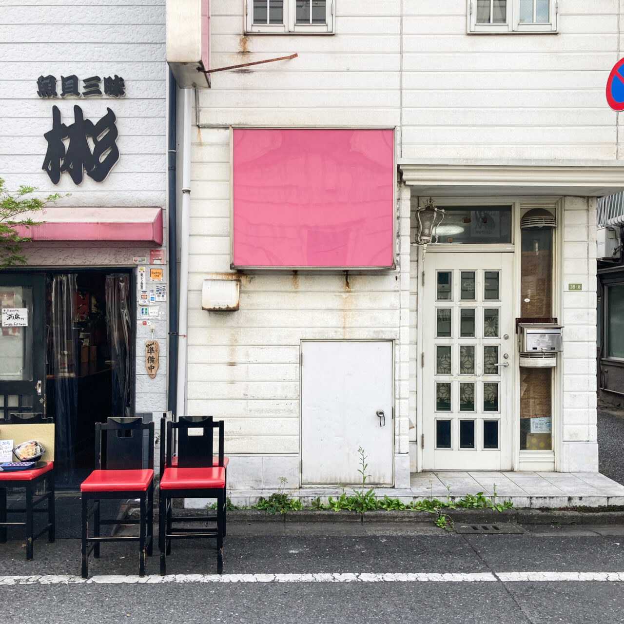 Photograph of a pink/faded red empty noticeboard on the side of a white panelled building, with the front door to the right set slightly back and in front 2 red and black chairs at the side of the road, in front of a restaurant next door.
