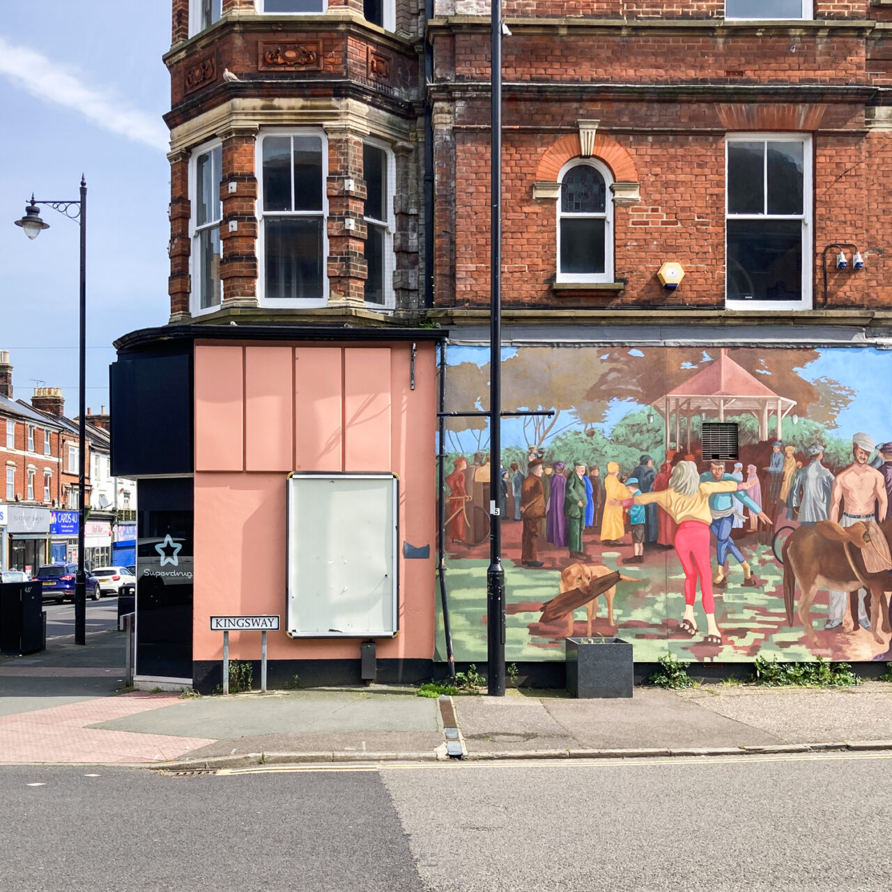 Photograph looking across a road to a wall that has a colourful painted mural to the right, and empty noticeboard/advertising board mid-left, a street sign saying 'KINGSWAY', windows in the brick wall above and street lamp and road visible to the left.