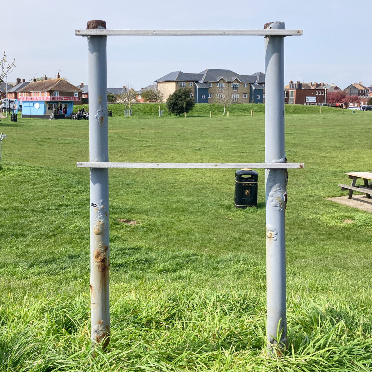 Photograph of a metal structure that probably previously held a notice. Behind is a grassy field with a bin and picnic table, a small cafe and houses are visible in the distance.