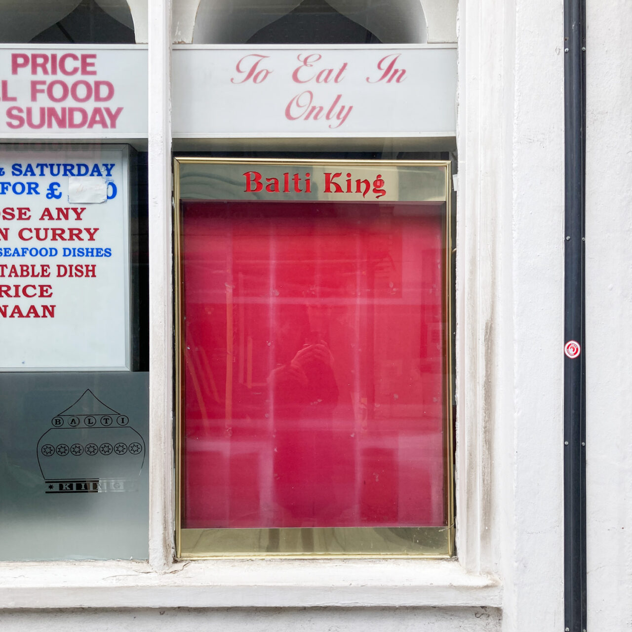 Photograph of a red display board with faded areas showing where paper had previously been displayed, with a gold frame that says 'Balti King' at the top. It sits in a window that has 'To Eat In Only' and parts of words relating to food and menus around it, with a black plastic pipe on the white wall to the right.