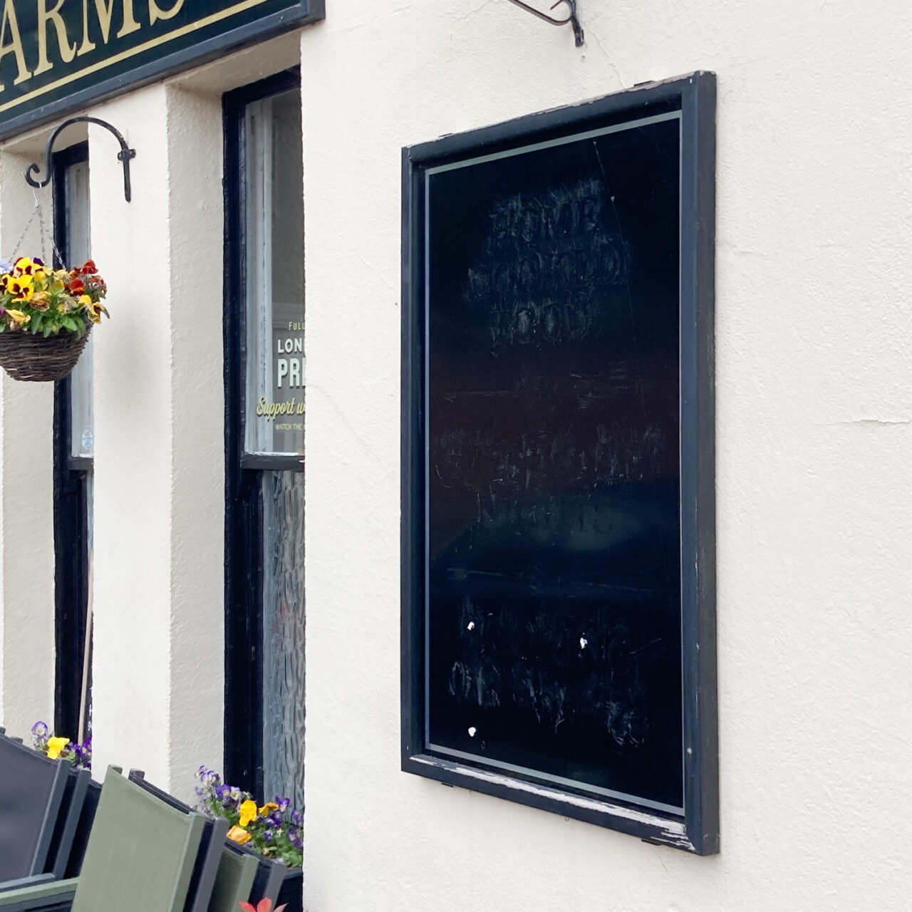 Photograph of a black menu board with remnants of writing barely visible, on a cream wall of a pub with windows and window box and word 'arms' partially visible to the left.