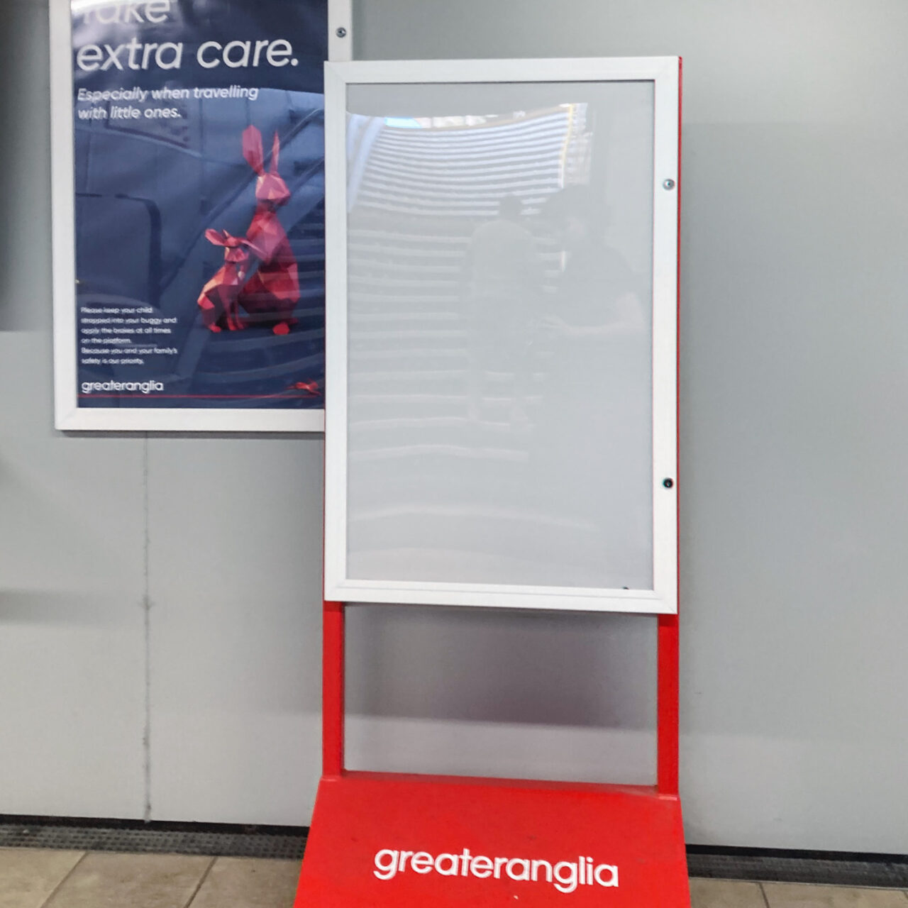 Photograph of a freestanding empty noticeboard with 'greateranglia' on the red sign at the bottom. Behind is a wall with a framed poster saying 'Take Extra care'.