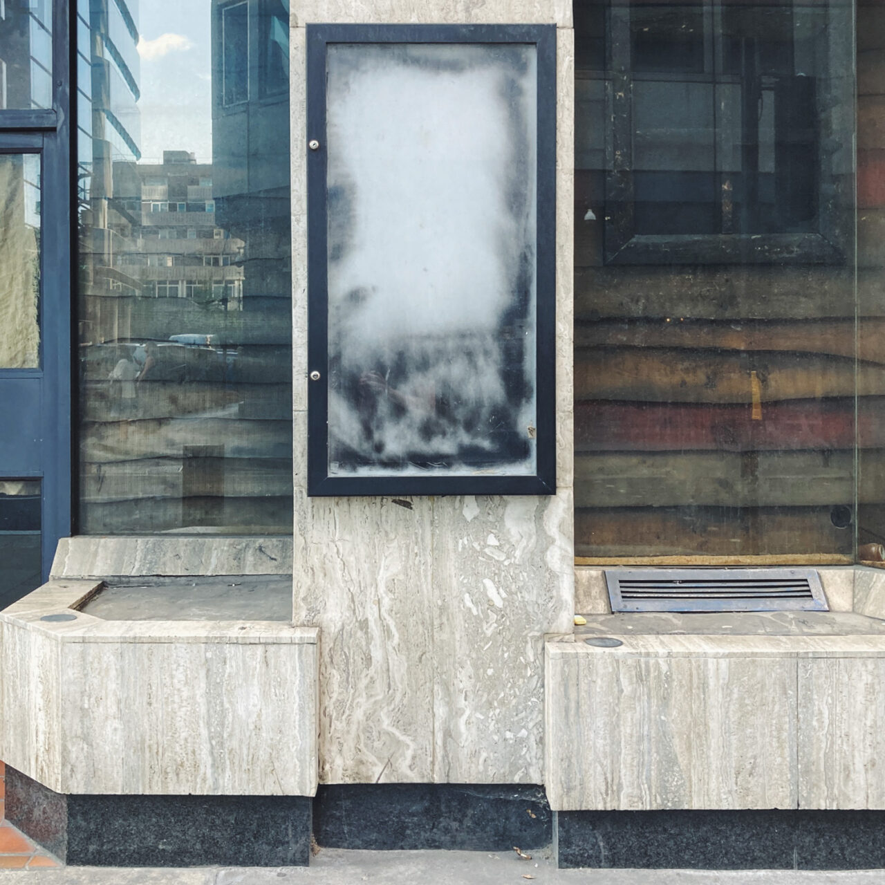 Photograph of an empty noticeboard on a pillar between two windows, with marble effect areas below.