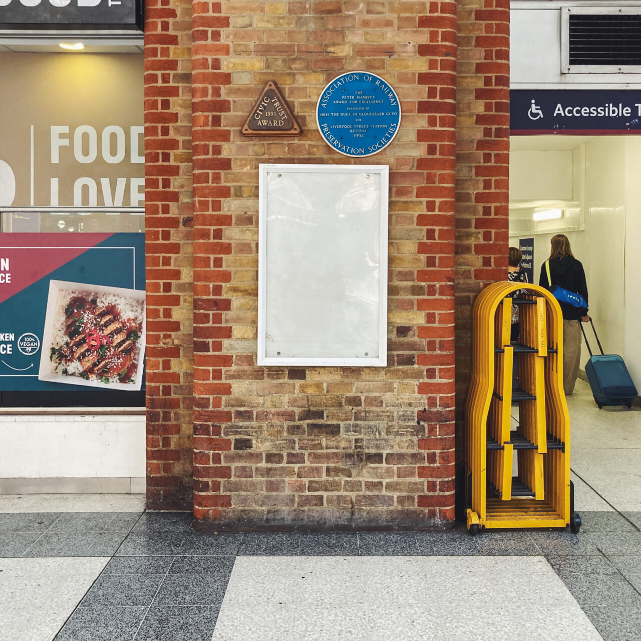 Photograph of an empty noticeboard on a brick wall, with blue plaque and triangular plack above, shop behind to the left and walkway with yellow retractable barrier to the right and tiled floor in front.