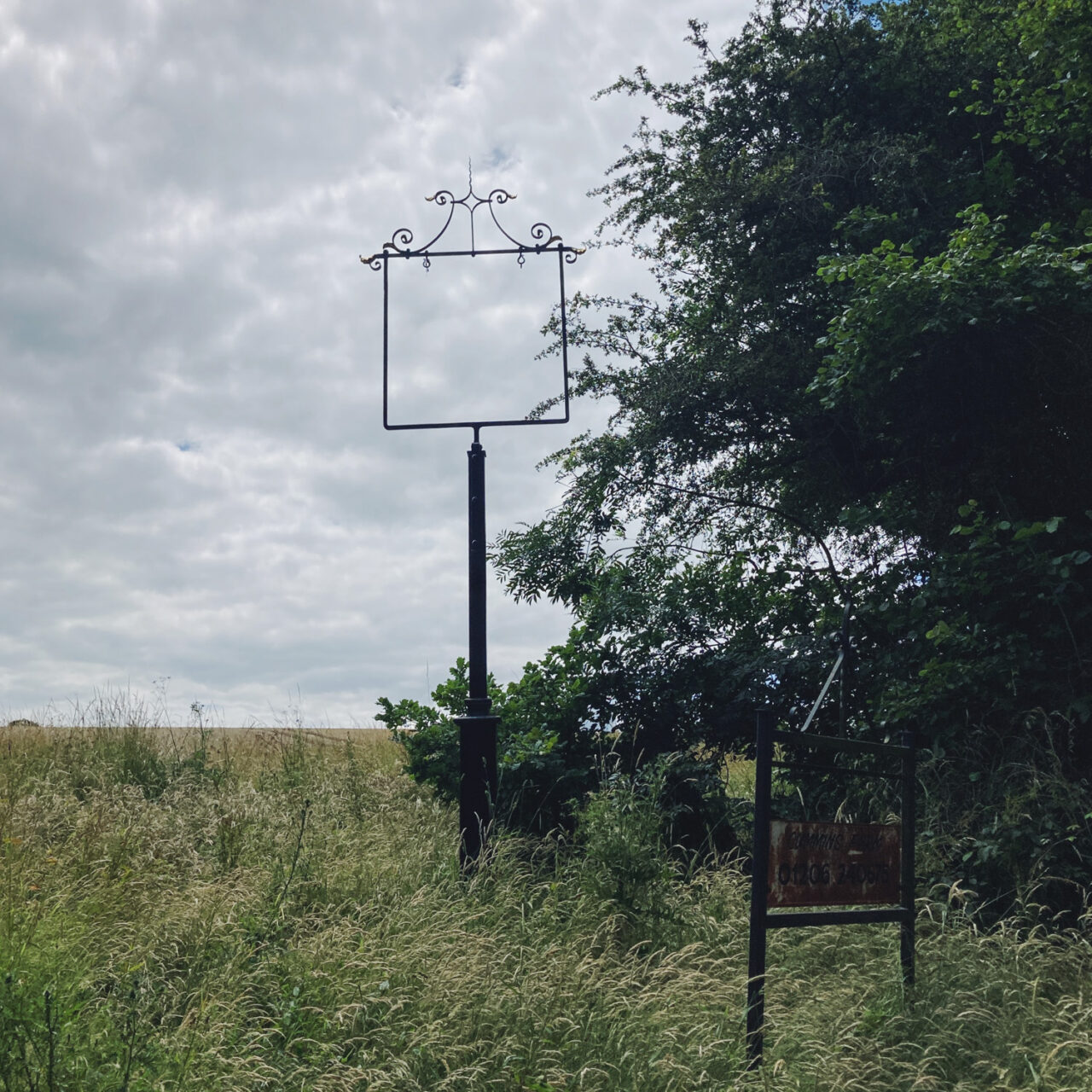 Photograh of a pub/town sign with the sign removed, with field below, cloudy sky above and trees to the right.
