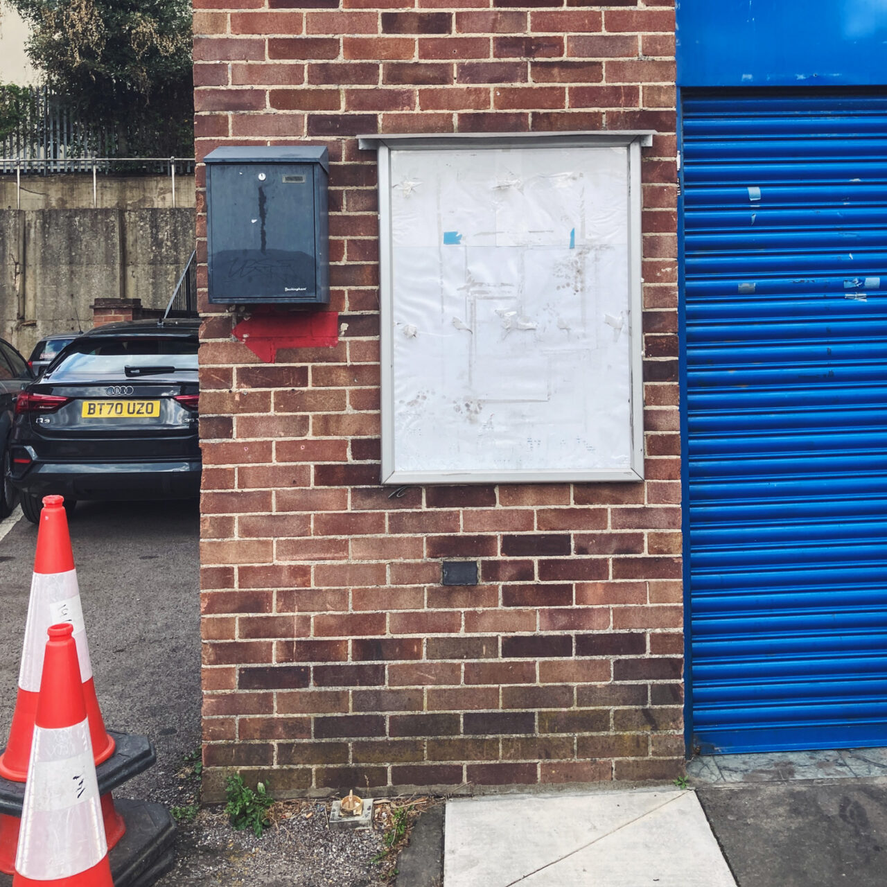 Photograph of an empty white noticeboard with remnants of tape, mounted on a brick wall with a grey metal box to the left, to the far left there are 2 traffic cones with a car behind and to the right a blue shutter.