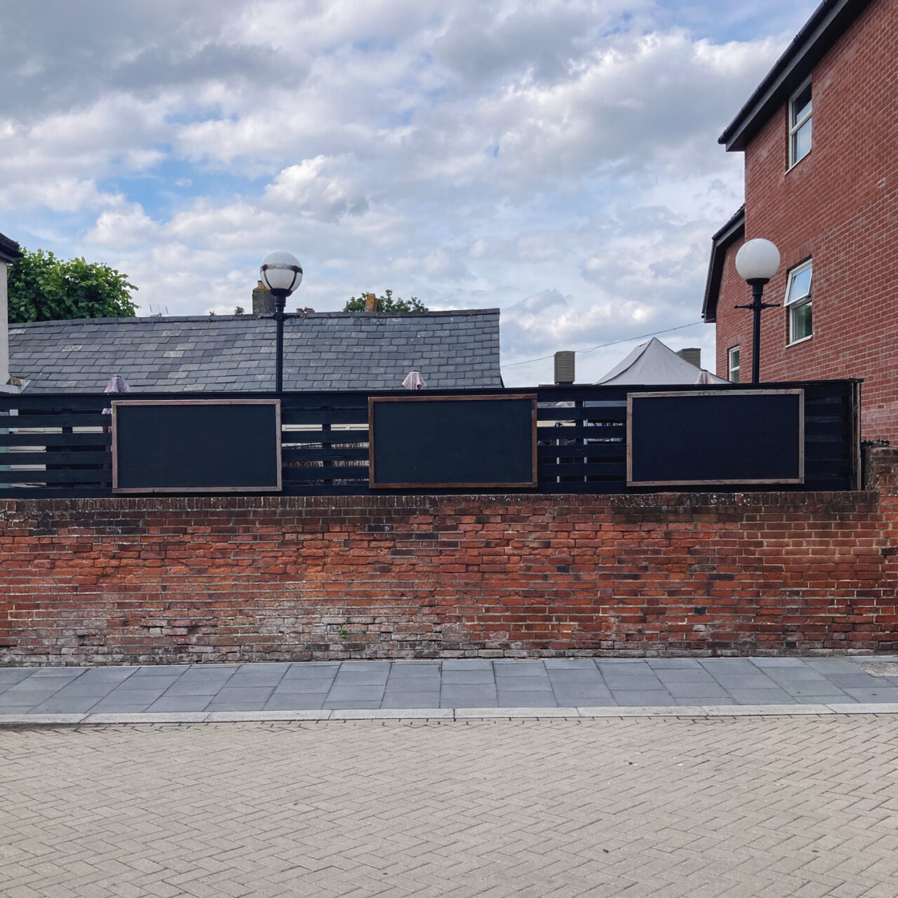 Photograph of 3 empty black noticeboards mounted on a black wooden fence above a brick wall, with a tiled floor in front and cloudy sky above.