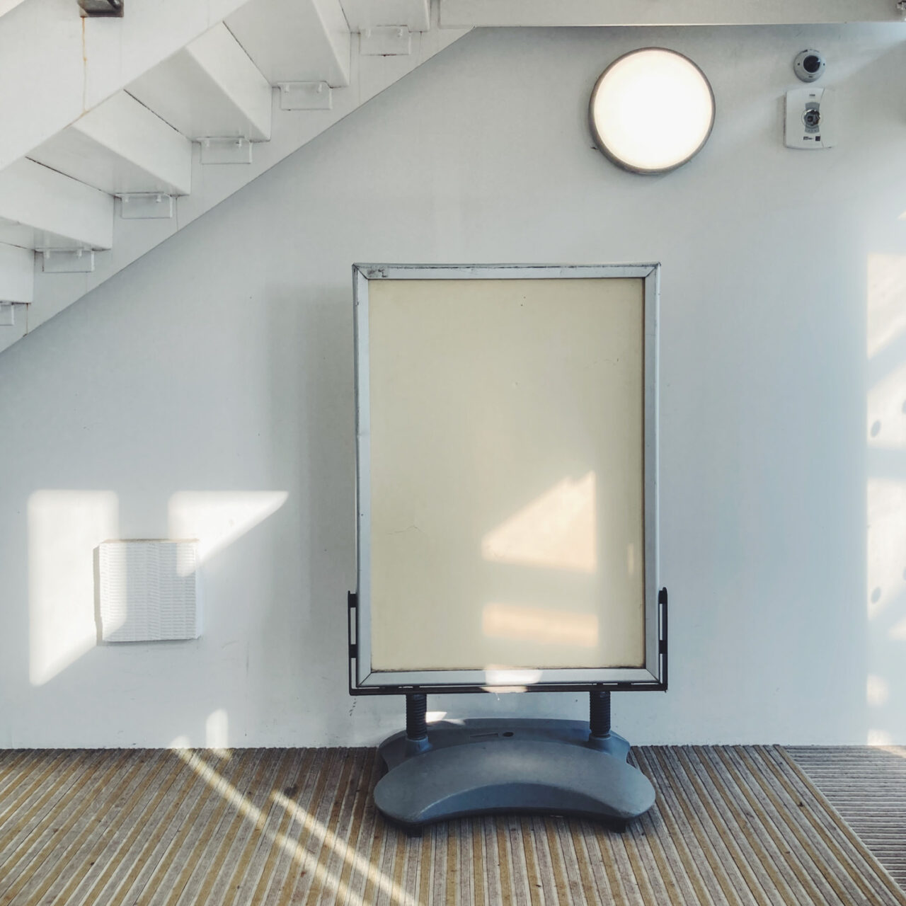 Photograph of an empty cream noticeboard on a plastic base, with white wall with a round light and staircase above, carpeted floor in front and bits of evening light casting triangular and line shapes across the scene.