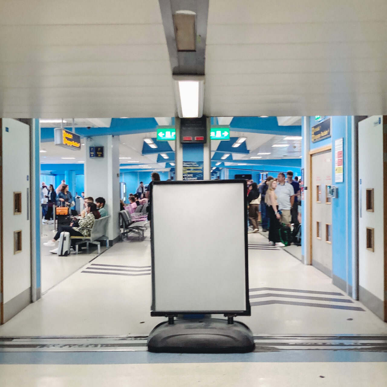 Photograph of an empty cream noticeboard on a plastic base in a corridor with an airport gate behind with people milling about.