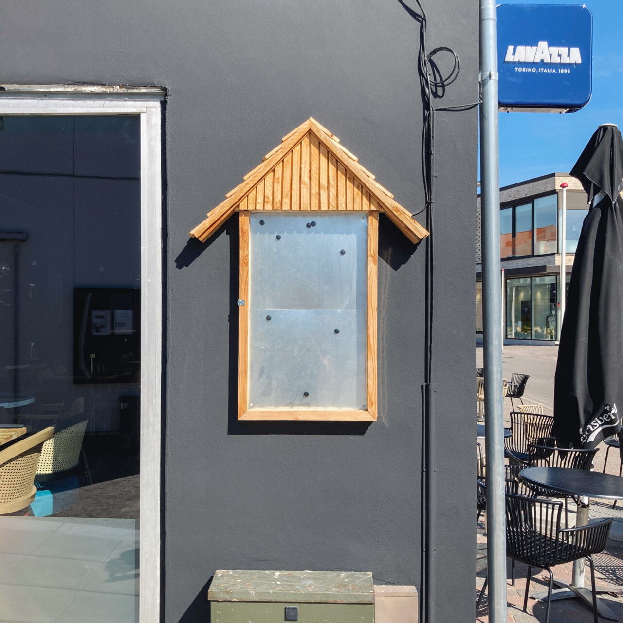 Photograph of a metal and wood empty noticeboard in the shape of the outline of a house, mounted on the side of a café painted with a dark grey wall, window to the left, seats, Lavazza sign and parasol to the right, with a khaki metal box below.