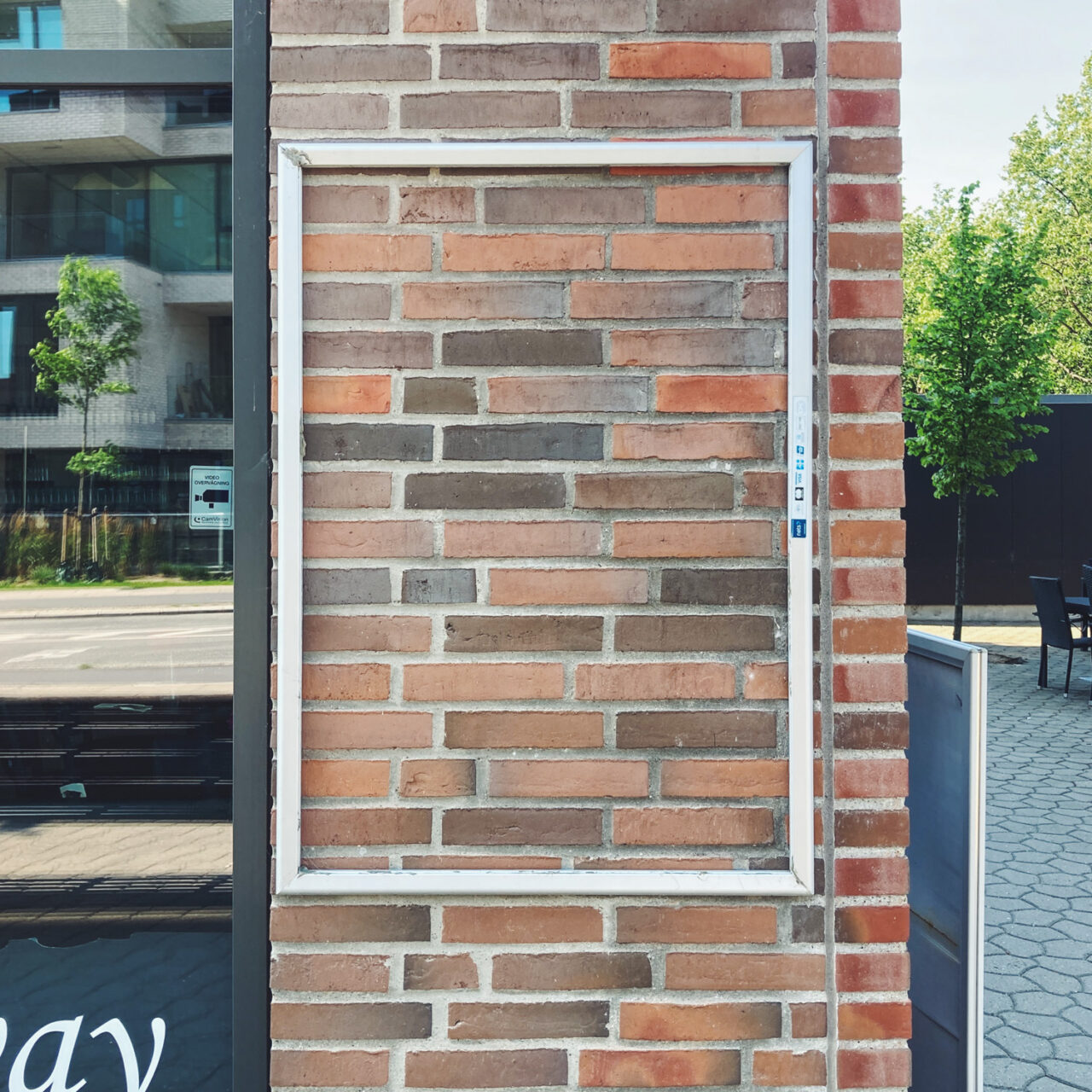 Photograph of an empty metal frame mounted on a brick wall with a window to the left and street to the right with a tree in the distance.