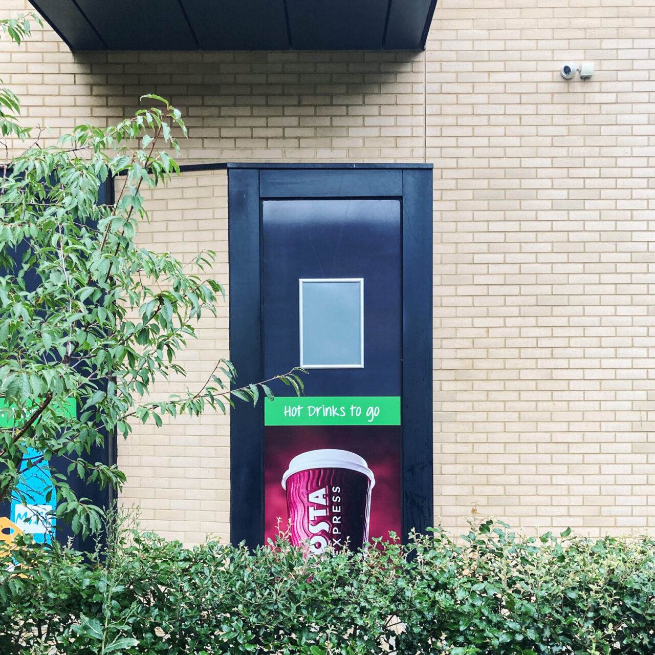 Photograph of a door with an empty noticeboard with a green sign saying 'HOT DRINKS TO GO' and a partially visibe Costa Coffee advert below, in a brick wall with a hedge and a tree below and to the left in the foreground.