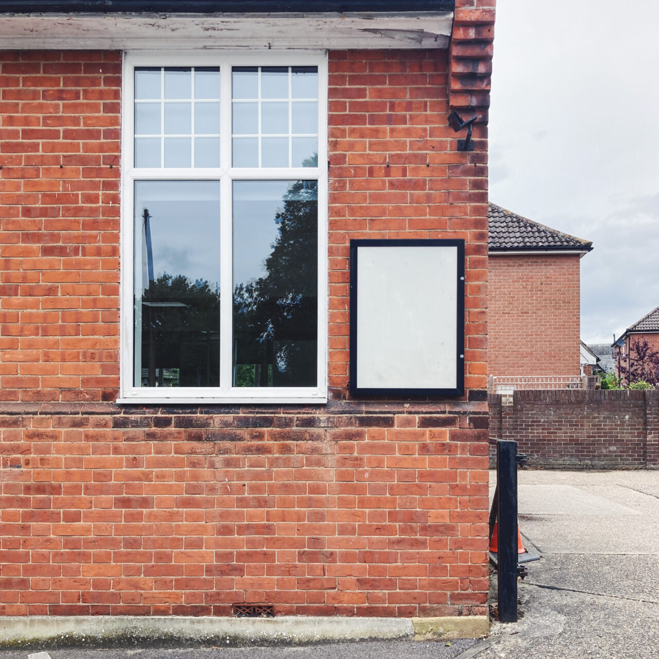 Photograph of an empty noticeboard with a black frame on a brick wall, with a window to the left of the frame and a bollard, carpark and building to the right and behind.