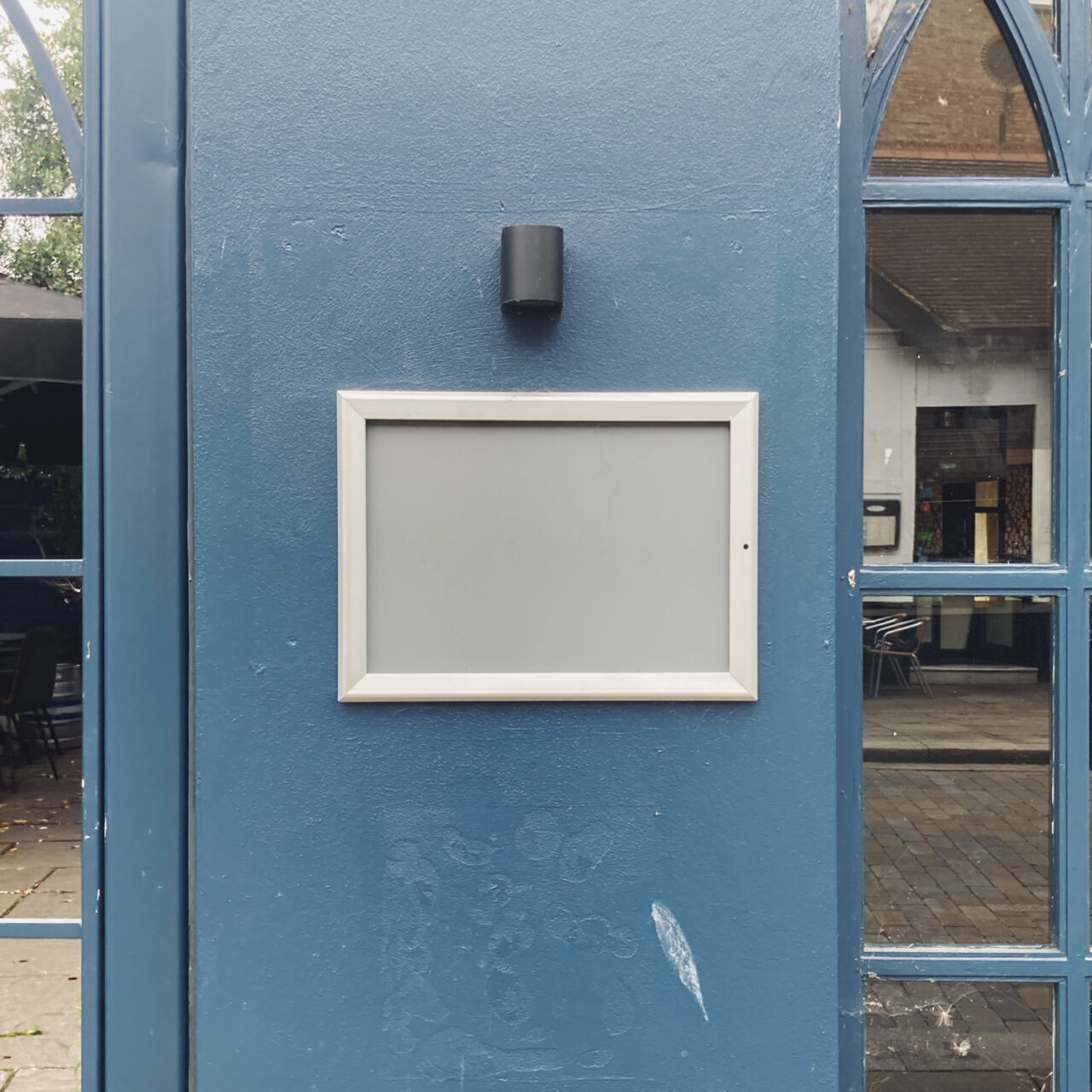 Photograph of an empty grey and metal framed noticeboard on a dark blue wall with mirrored glass panels either side.