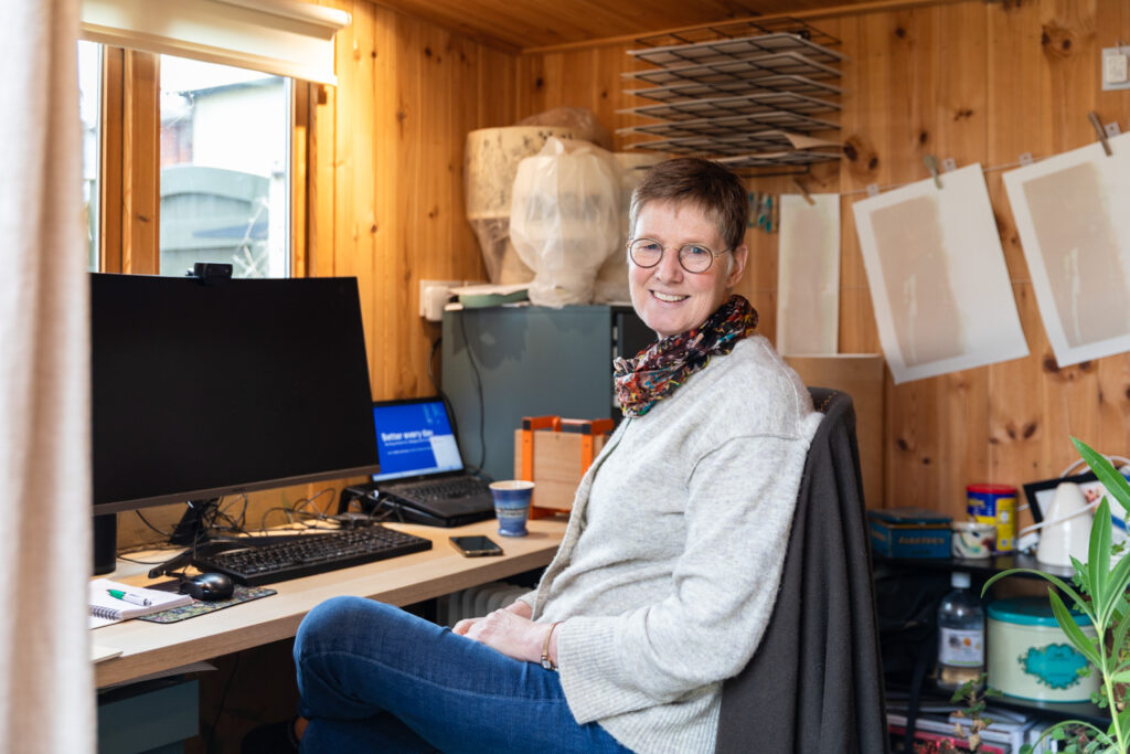 Photograph of Nel Mooy sitting at her desk with computers and print equipment visible behind. She is smiling into the camera and wearing a grey woolen top, blue jeans and a colourful scarf.