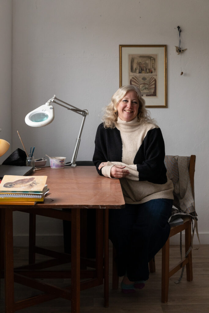Photograph of artist Madi Boyd sitting at a wooden desk in her studio. She is smiling into the camera and wearing a black and cream jumper. Behind her is a framed print and ornament hanging on a white wall and on the desk are notebooks, materials and a lamp with built in magnifier.