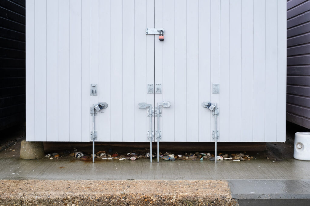 Clacton-on-Sea, UK, February 2025. Photograph of the bottom of a beach hut, with long bolts connecting to the concrete floor below. Under the beach hut is a collection of stones, many of them painted.