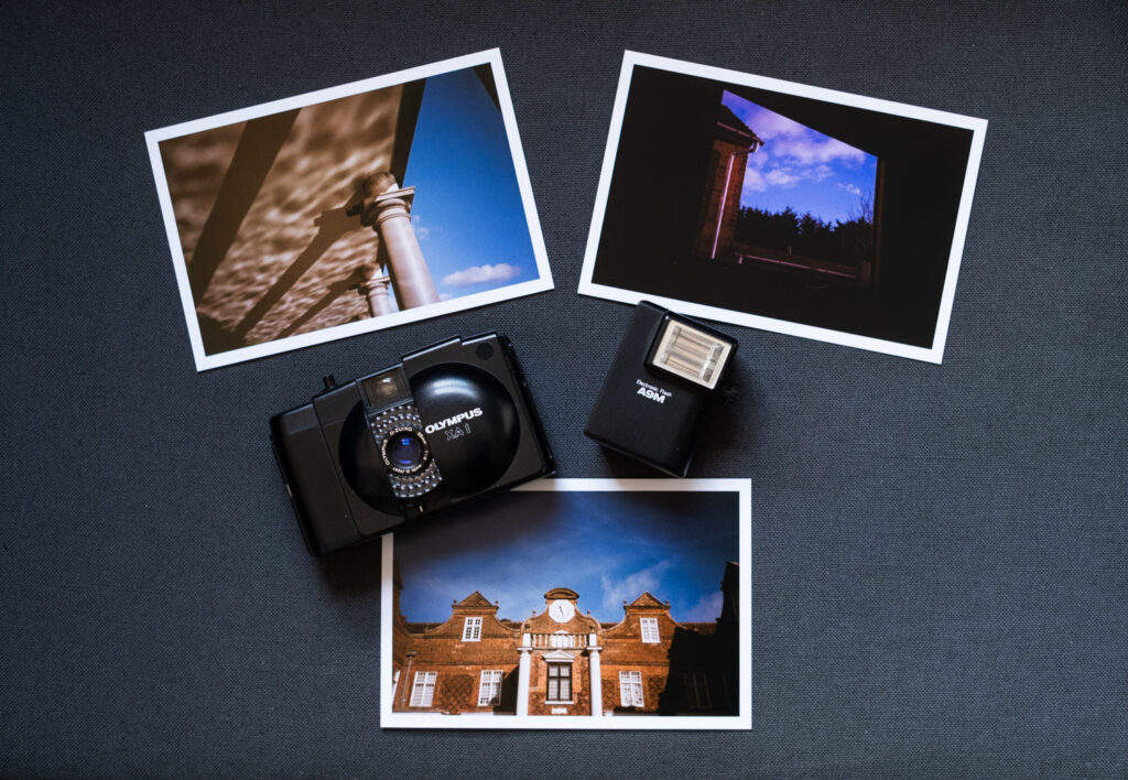 Photograph of an Olympus XA 1 camera with an A9M flash, on a dark grey fabric background with 2 prints above and one below. The prints show (clockwise from top left) reflections of water on a ceiling supported by columns and a blue sky with a cloud to the right; a view through a window of part of a house, tops of trees and blue sky with clouds above; part of the facade of Christchurch Mansion in Ipswich, showing a brick building with a clock in the top centre and blue sky and clouds above.