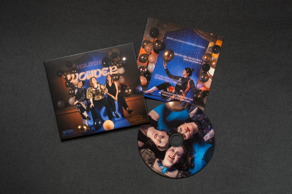 'Hold the Wonder' by Wise Woman. Photograph by Jayne Lloyd. Photograph of a CD and packaging for Hold the Wonder by Wise Woman, photographed on a dark grey fabric background. The EP case is to the left, with text reading 'HOLD THE WONDER, wise woman, LIVE at Blaxhall Village Hall' and a photograph of 3 women sitting on the edge of a stage with a strip of blue background running through the centre and balloons all around them. The CD to the lower right of the image shows a photograph of the faces of the 3 women lying on a blue background in a circular formation, and the EP insert is above that, with a photograph of oen woman sitting sideways on, holding a gold balloon, with a blue background lit with fairy lights and surrounded by balloons behind.