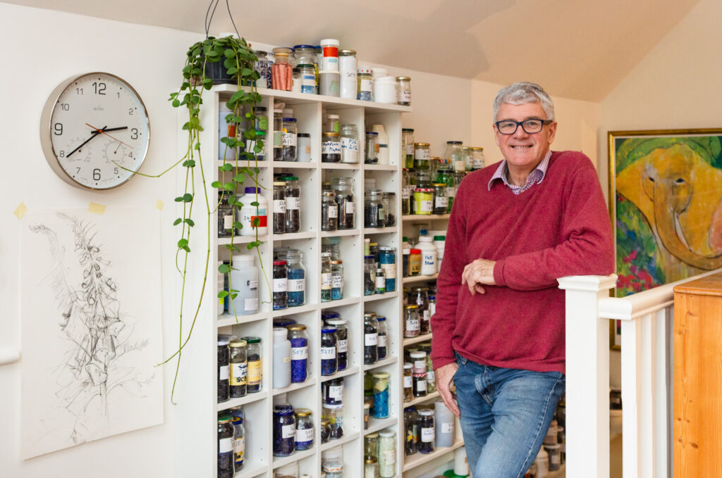 Photograph of Fred Rich, wearing a cerise jumper, blue jeans and glasses, leaning on a bannister, with white shelving to his right holding jars and pots full of materials. To the left of the frame is a clock, a houseplant and a drawing of a hollyhock taped to the wall, and a painting of an elephant can be seen behind Fred to the right of the frame.