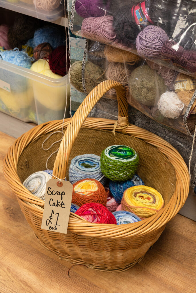 Photograph of a wicker basket on a wooden floor, filled with multicoloured balls of wood, with 'Scrap Cake £1 each' handwritten on a paper label, with boxes of wool visible in the background.