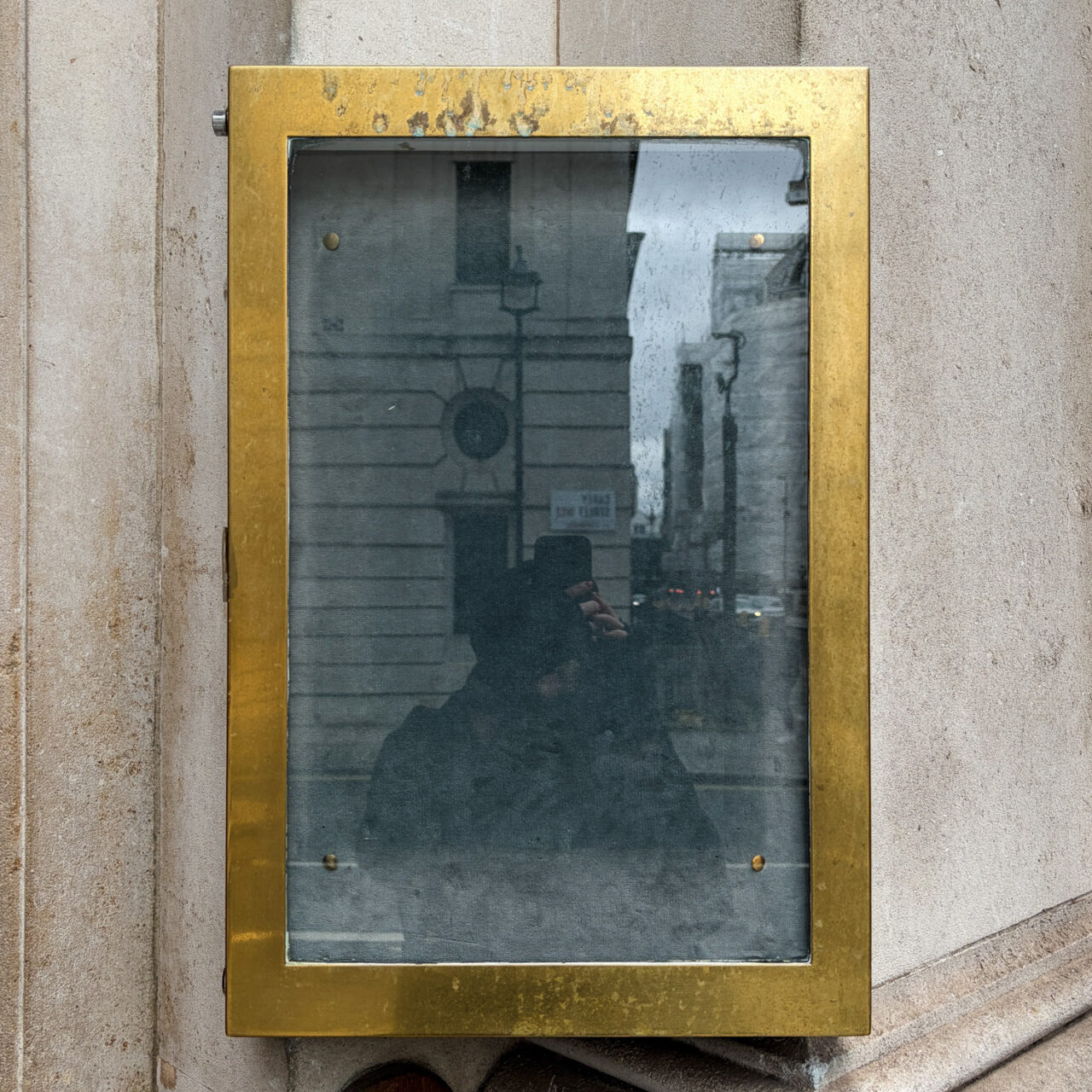 Photograph of an empty brass frame on a wall, with buildings and a person holding a phone up reflected in the glass.
