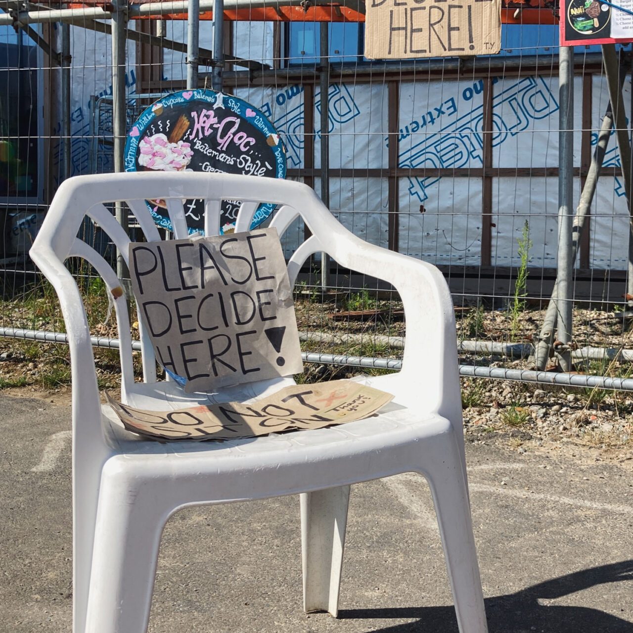 Photograph of a plastic garden chair with with a handwritten sign saying'PLEASE DECIDE HERE!', behind it is scafolding, another cardboard sign with the same message, and adverts for hot-choc and icecreams.