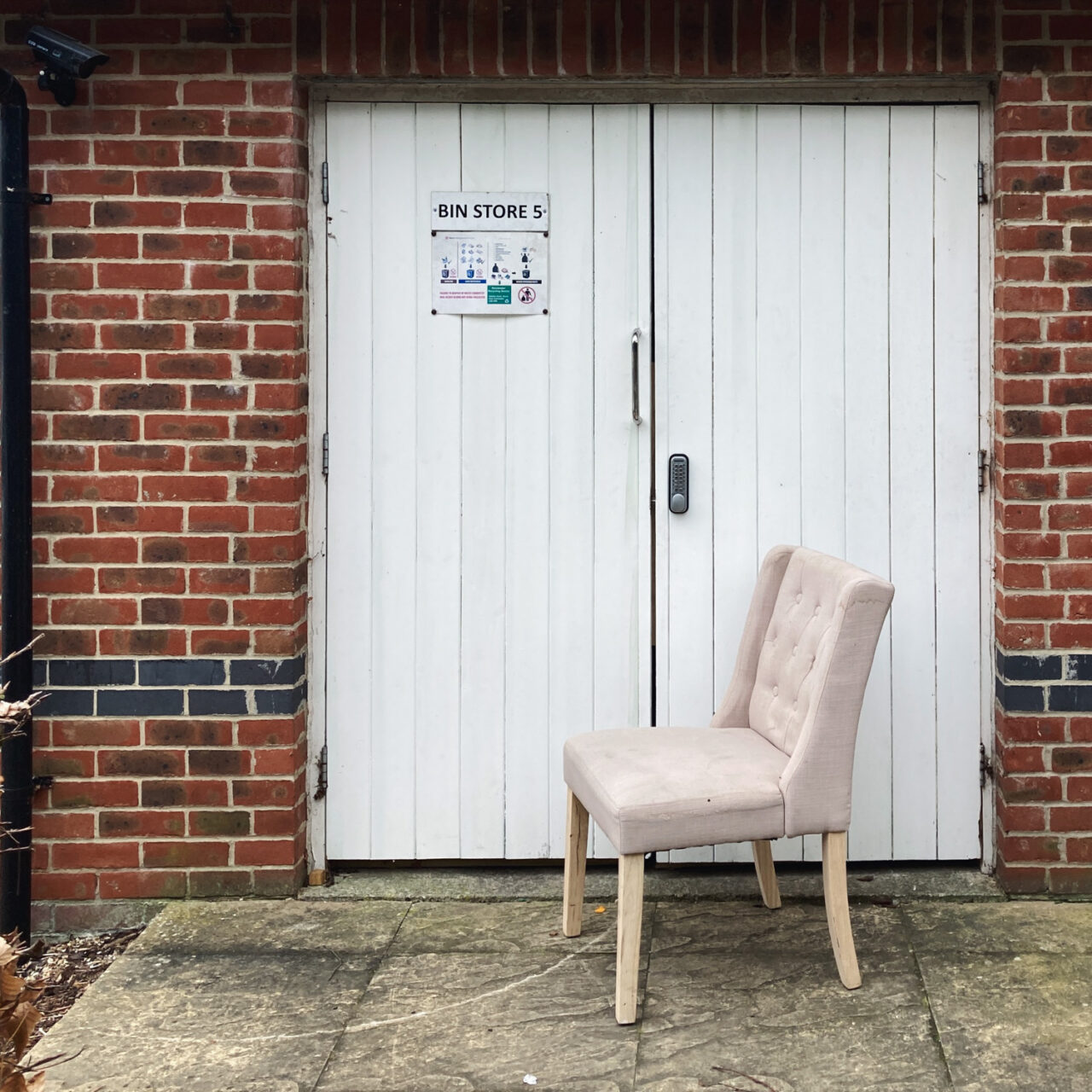 Photograph of a beige upholstered dining chair in front of a brick building in a housing estate with white wooden doors and a sign saying 'bin stores' to the left.
