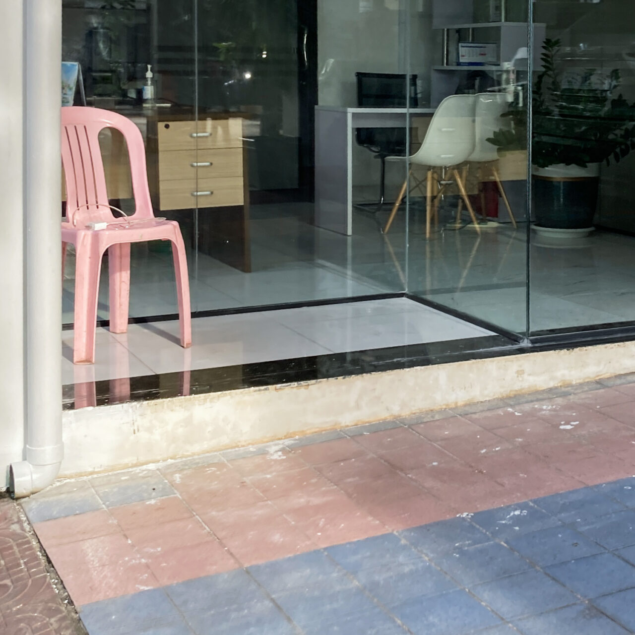 Photograph of a pink plastic garden chair in the entrance of a glass-fronted office, with pink and blue tiles on what appears to be a driveway in the foreground.