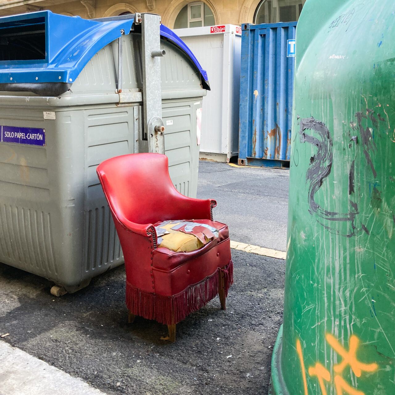 Photograph of a red armchair with a seat that is ripped with the foam visible, it appears to have been fixed with tape many times in the past. The chair is on the roadside between bins and bottle recycling facilities, with ornate buildings visible on the opposite side of the road.