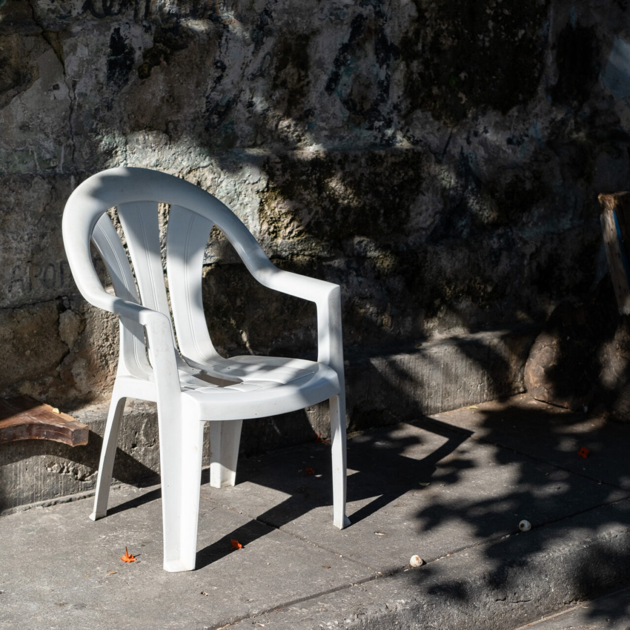 Photograph of a white plastic garden chair on a pavement, shadows of foliage to the right and orange flowers in the road in front.