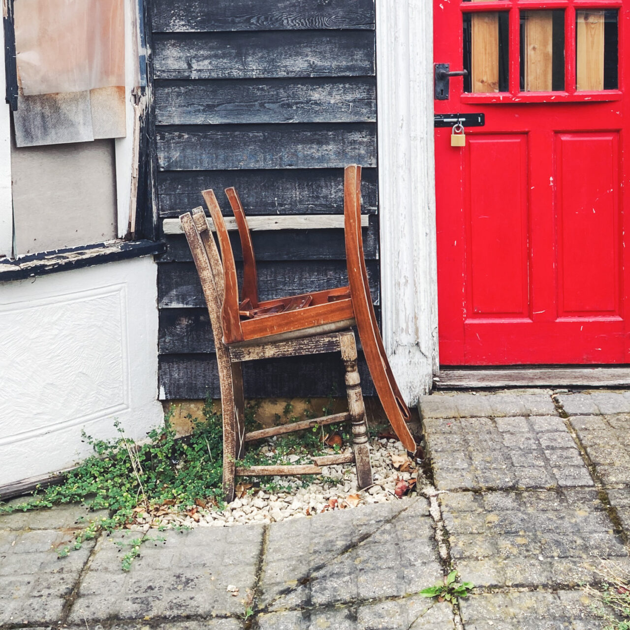 Photograph of two chairs, one upside down on top of the other, in front of a building with red door to the left, boarded up bag window to the left and paving slabs in the foreground.