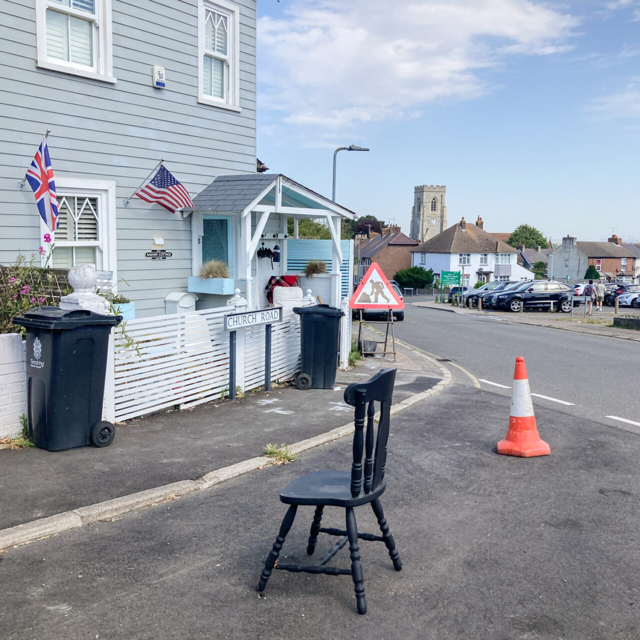 Photograph of a black wooden kitchen chair in the road outside a house, with traffic cone to the right behind. The house has a light blue wooden facade, British and US flags flying and black wheely bins. In the distance is a construction work sign, a car park and buildings including a church.