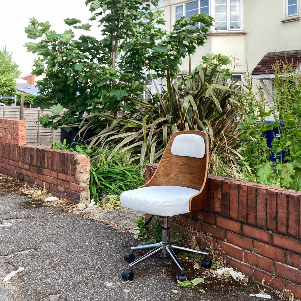 Photograph of a wood-backed office chair on a pavement with a red brick wall, garden and house behind.